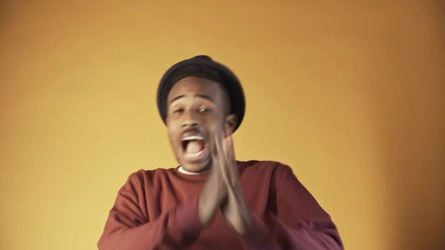 Portrait shot of emotional black man in hat covering eyes with his hands, then laughing and screaming at camera while posing isolated on orange background