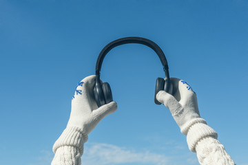 Female hands in the winter mittens with a headphones on the cloudy blue sky. Concept
