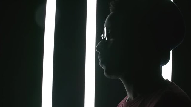 Side view portrait shot of silhouette of black man in round glasses standing in dark studio; fluorescent tubes swinging behind him