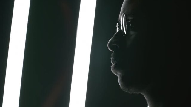Side portrait with close up of face of black man in round glasses and hat standing in dark studio with fluorescent tubes swinging behind him