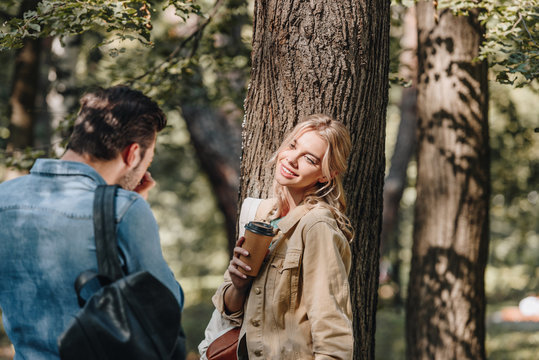 man taking picture of smiling girlfriend with coffee to go in park