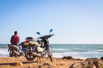 A guy in a burgundy waistcoat on a motorcycle against the background of the ocean. The coast of the Atlantic Ocean. The beach is near Agadir. Africa Morocco