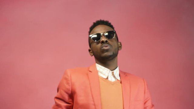 Portrait shot of stylish black man in sunglasses and salmon colored standing against bright pink background and rapping before camera