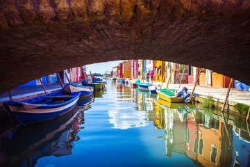 Fototapeta na wymiar View under bridge on typical street scene showing brightly painted houses and boats with reflection along canal at Islands of Burano in Venice, Italy