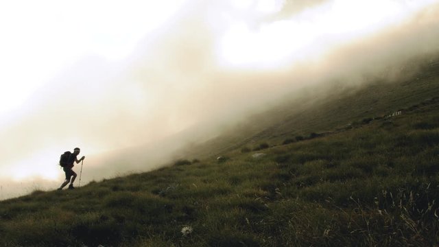 Young trekker man walking up the mountain in the fog with backpack, boots and trekking poles. Clouds and myst moving on the background. Abruzzo Italy.
