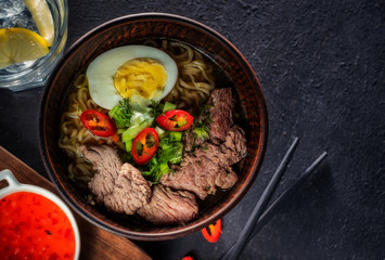 Asian noodle soup with beef, egg, red pepper and herbs on a dark table, top view