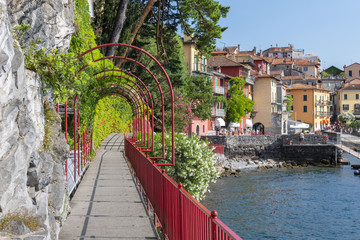 Fototapeta na wymiar Red garden arch (Walk of Lovers) on coastline leading towards the beautiful and historic city of Varenna on the edge of Lake Como in the northern Italy.