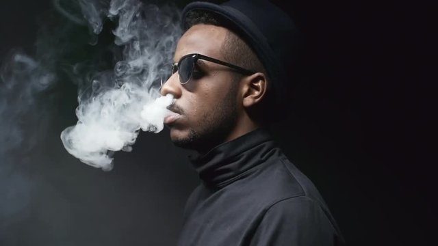 Panning side view portrait shot of cool black man in sunglasses and hat standing in dark studio and exhaling smoke from his mouth and nose, then looking at camera and talking