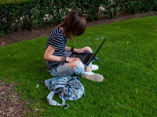 A teenage woman studying with a laptop sitting on the grass in a park of a university in a university campus
