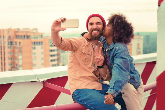 Taking selfies. Positive bearded young man smiling and taking photos while his beautiful girlfriend kissing him