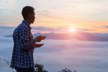 Fototapeta premium A tourist holding a cup of coffee on the mountain,The valley is covered with fog and has a beautiful sunrise. Beauty of nature, tourism and traveling concept.