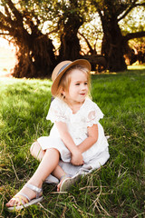 A cute baby girl in a straw hat and white dress is sitting on the grass.