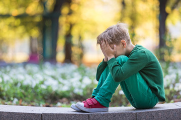 Fototapeta premium Caucasian boy worries because of problems. Sits one summer in the park having hidden the face in hands and cry