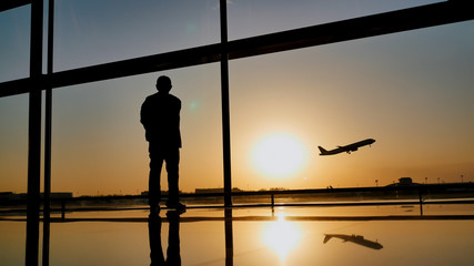 Silhouette of a tourist guy watching the take-off of the plane standing at the airport window at sunset in the evening. Travel concept, people in the airport. © Довидович Михаил