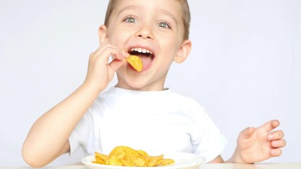 A child with pleasure eating potato chips sitting at a table on a white background. The boy is eating chips and smiling.