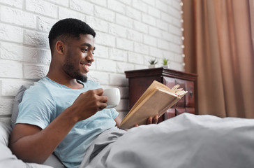 Black man drinking coffee and reading book in bed