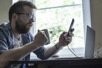 A thoughtful bearded man with glasses is holding a smartphone and a cup with a hot drink in his hands, on the desk is a laptop, in the background of a window and a flower, in the early morning.