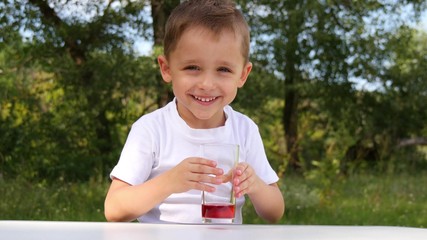 A child with joy drinks berry juice from a glass glass on a background of nature.