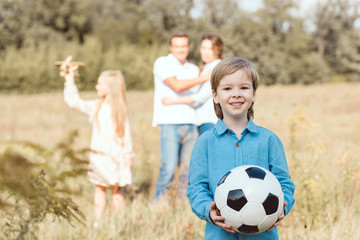 adorable little kid holding ball while his family standing blurred on background in field