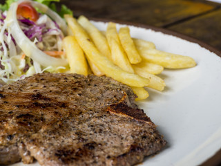 close up of T-bone steak with salt and pepper, golden french fries and green vegetables. on white plate, Fork and knife, wooden table in the restaurant