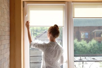 Young woman opening roller curtains at home