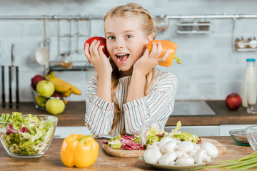 happy little child with bell peppers looking at camera while making salad at kitchen