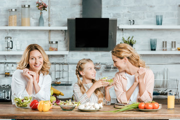 happy little child preparing salad with mother and grandmother at kitchen