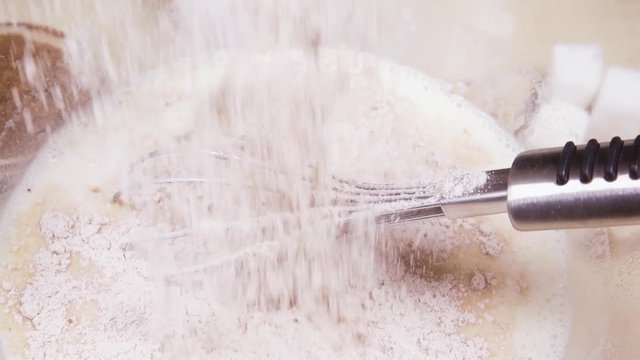 Slow motion pour the cocoa flour into a glass bowl with milk mixture and a metal whisk close up