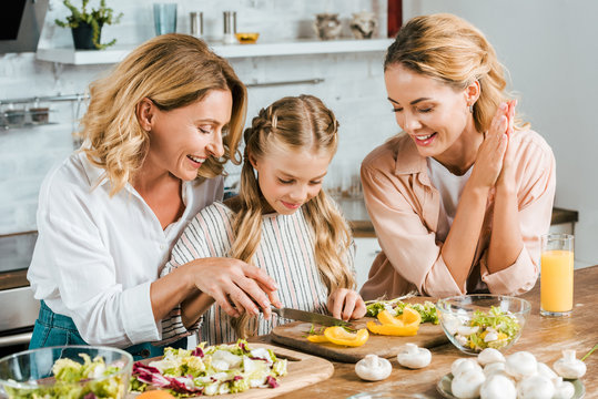 Adorable Little Child Cutting Vegetables For Salad With Mother And Grandmother At Home