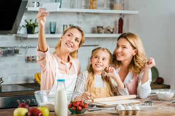 beautiful young woman taking selfie with her mother and daughter while cooking at home