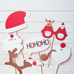 A Christmas props for a photo booth (a Santa Claus hat, his beard or mustache, funny glasses, golden deer antlers, a smoking pipe, the phrase "hohoho") is on a white wooden background.  Flat lay