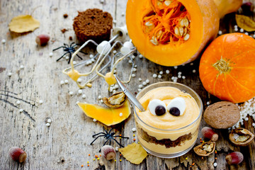 Halloween monster dessert in glass, layered dessert of pumpkin mousse with chocolate cookie crumb with eyes on a wooden table with fresh pumpkins