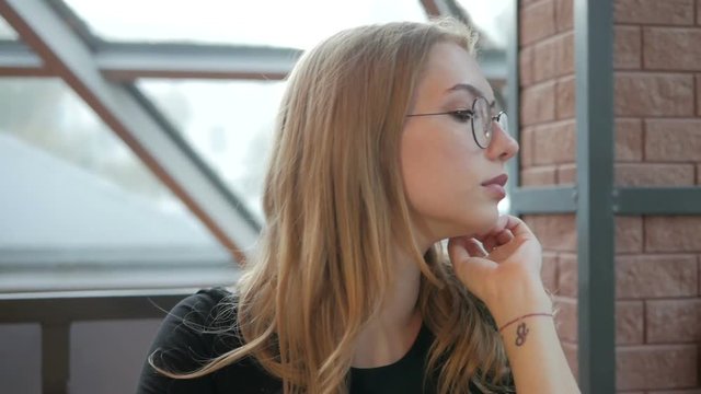 Young beautiful girl in glasses sitting in a cafe by the window looking at the sides