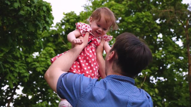 Dad plays with little daughter, throws up baby with his hands, child is happy and laughs. Dad is holding small daughter in summer park.