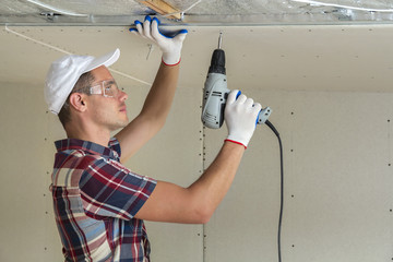 Young man in goggles fixing drywall suspended ceiling to metal frame using electrical screwdriver...