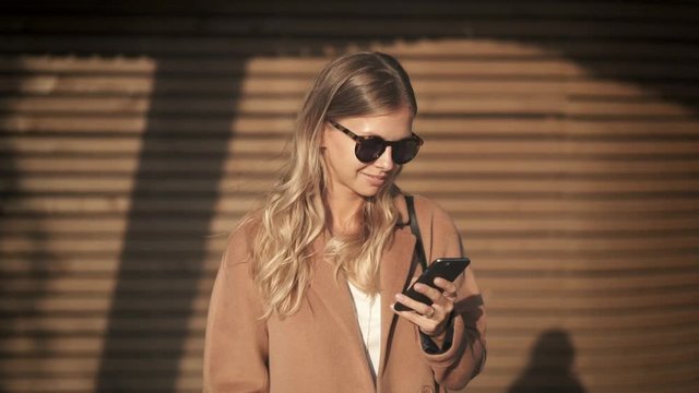 Attractive young woman wearing a coat and glasses is standing in autumn street and looking at her smartphone, smiling. Social media and street fashion concept. Slider slow motion portrait shot.