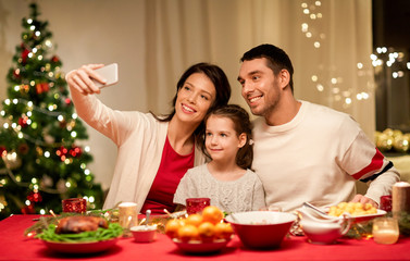 holidays, family and celebration concept - happy mother, father and little daughter having christmas dinner and taking selfie picture by smartphone at home