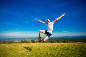 Fototapeta premium Summer hiking in mountains. Young tourist man in cap with hands up on top of mountains admires nature. Travel concept