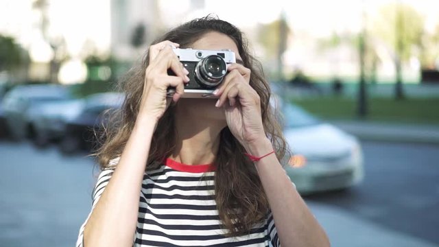 Smiling young photographer with long curly hair wearing a striped shirt is taking a shot with a vintage camera in downtown. Slider real time medium shot