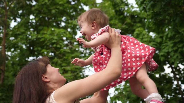 mom plays with little daughter, throws up baby with his hands, child is happy and laughs. little girl plays in hands of mother in summer park.