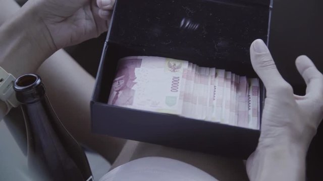 Caucasian woman sits in passenger seat inside of riding car and holds open small black rectangular box full of foreign eastern currency.