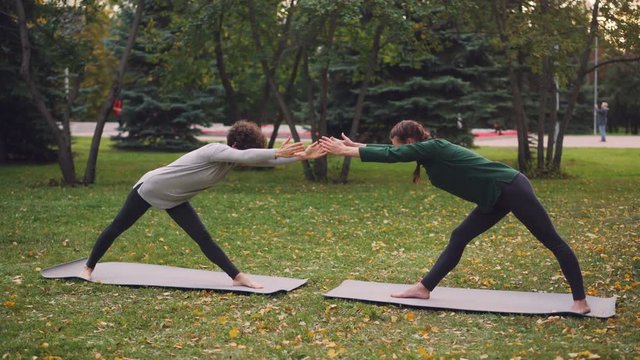 Slim young ladies are training outdoors in park doing hatha yoga together during pair practice and breathing fresh air. Beautiful autumn nature and people are visible.