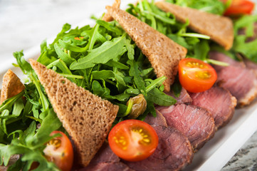 Salad from arugula and tomatoes. Slices of chopped black bread and sliced meat. Close-up on a white plate