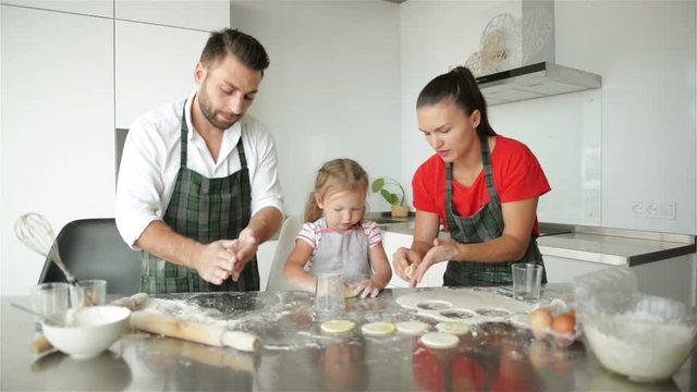 Cute Little Girl And Her Beautiful Parents Are Cooking. They Have A Lot of Fun Together And Smiling In Kitchen At Home.