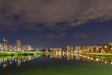 Fototapeta na wymiar Night cityscape of residential buildings and park with a pond in the urban outskirts