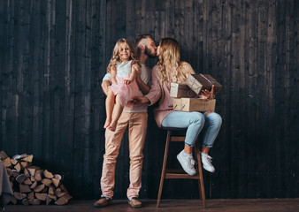 Family concept. Handsome father holds daughter on hands and kiss his wife with gifts in empty room against a wooden wall.