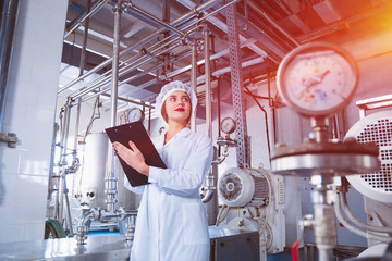 A young beautiful girl in white overalls makes notes in a tablet on the background of equipment of...