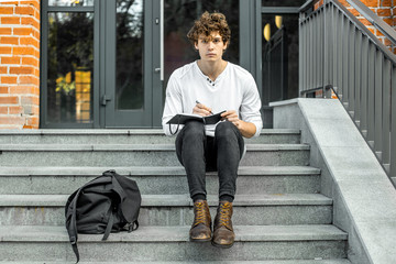 Pensive student sitting on modern building steps and making notes in notepad.