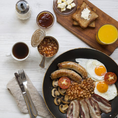 Full English breakfast in a pan with fried eggs, bacon, beans, sausages  and toasts on white wooden table, overhead view. Close-up.