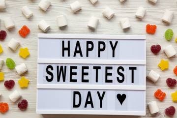 Modern board with text 'Happy Sweetest Day' word and candy over white wooden background, overhead view. From above, flat lay, top view.